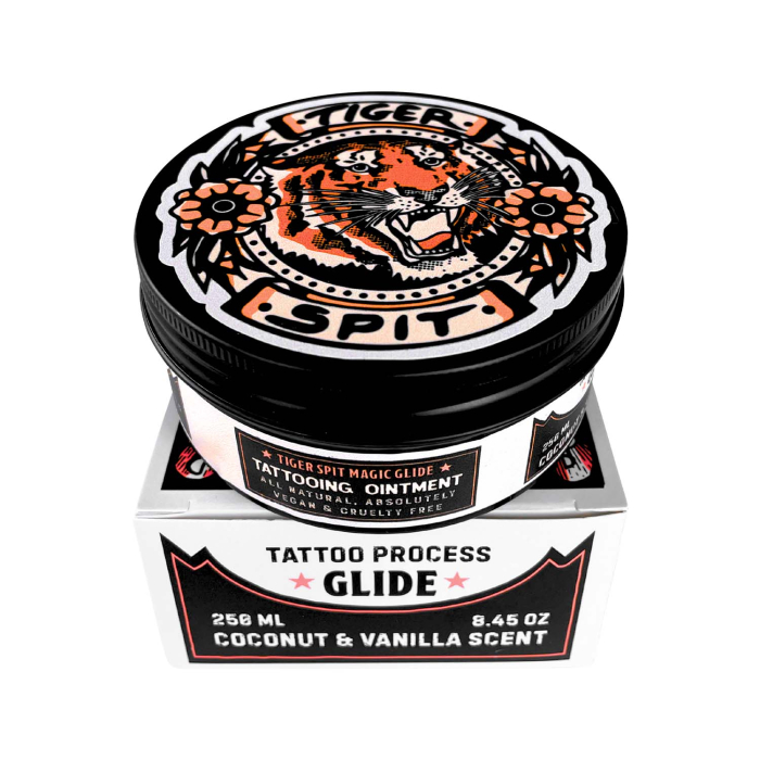 Un récipient rond et noir étiqueté « Tiger Spit Glide 250 ml », avec une illustration de tigre sur le couvercle, est posé sur une boîte assortie portant la mention « Tattoo Process Glide, Coconut & Vanilla Scent ».